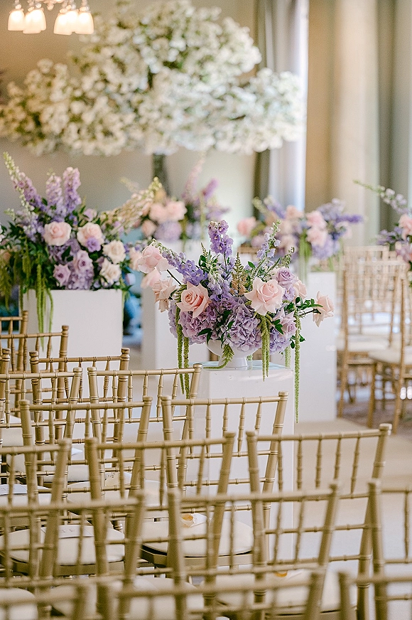 Ceremony floral decor with wedding ceremony flowers in pastel roses and hydrangea on white pedestals beside gold chiavari chairs indoors