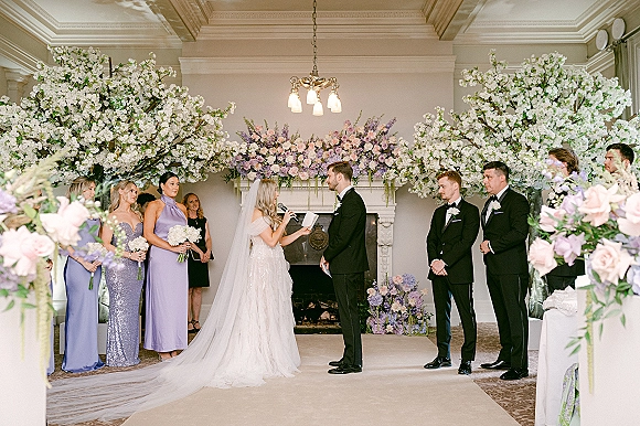 Wedding vows as bride reads from a vow book into a microphone, groom listening beside pastel floral arch and fireplace mantel backdrop