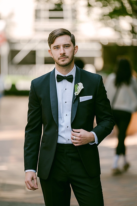 Groom portrait in a black tuxedo groom look with bow tie, boutonniere and pocket square, standing on an outdoor walkway with trees