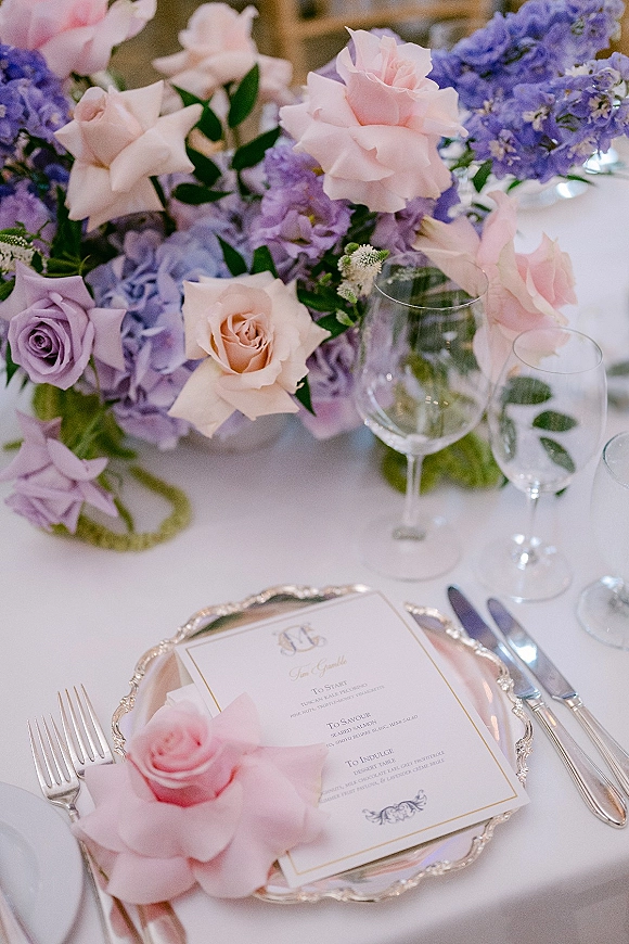 Reception tablescape with pastel wedding tablescape florals—pink and lavender roses, purple blooms, greenery, silver chargers, menu card, and wine glasses