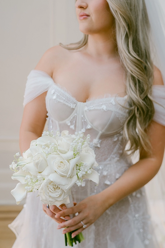 Bridal portrait of a bride in an off the shoulder wedding dress holding a white rose bouquet before a neutral indoor wall