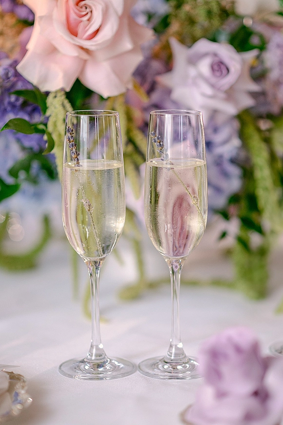 Champagne toast glasses with wedding champagne flutes filled with sparkling wine, garnished with lavender sprigs beside pastel florals on a white tablecloth