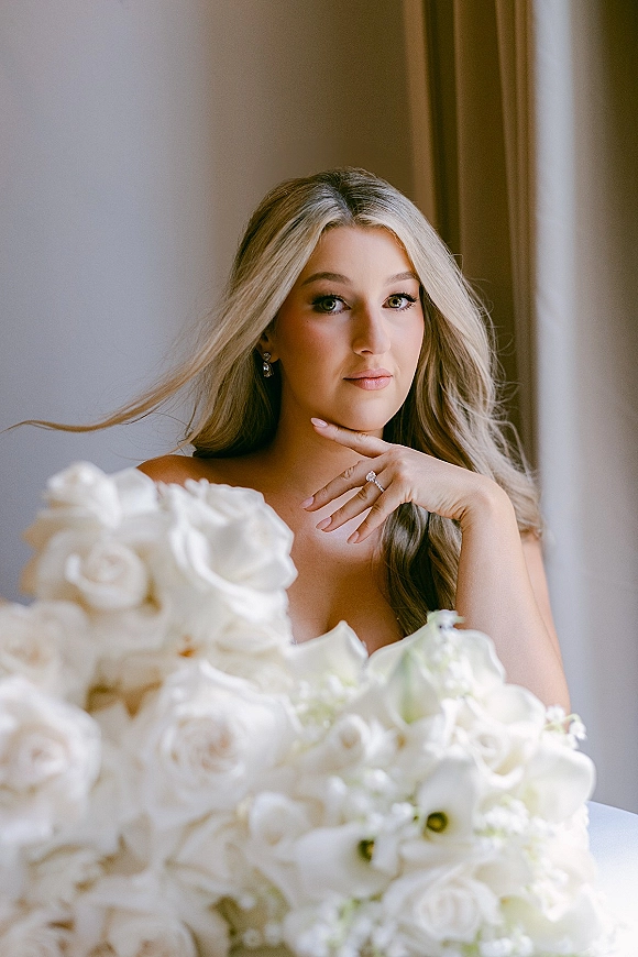 Bridal portrait with soft glam bridal makeup look, loose blonde waves, holding white rose bouquet by a window with sheer curtains