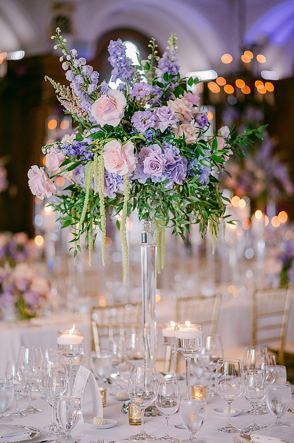 Reception tablescape with tall floral centerpiece of pink roses, purple blooms and hanging amaranthus, candles and glassware under string lights
