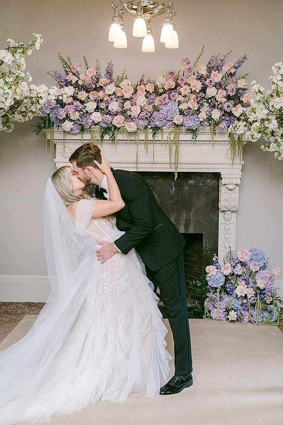 Wedding kiss portrait of bride and groom kissing, her veil flowing, framed by a chandelier and pastel floral fireplace mantel backdrop