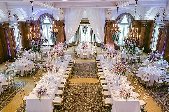 Reception tablescape with long banquet table setup, white linens, gold chiavari chairs, pastel florals, and candlelit centerpieces in a ballroom