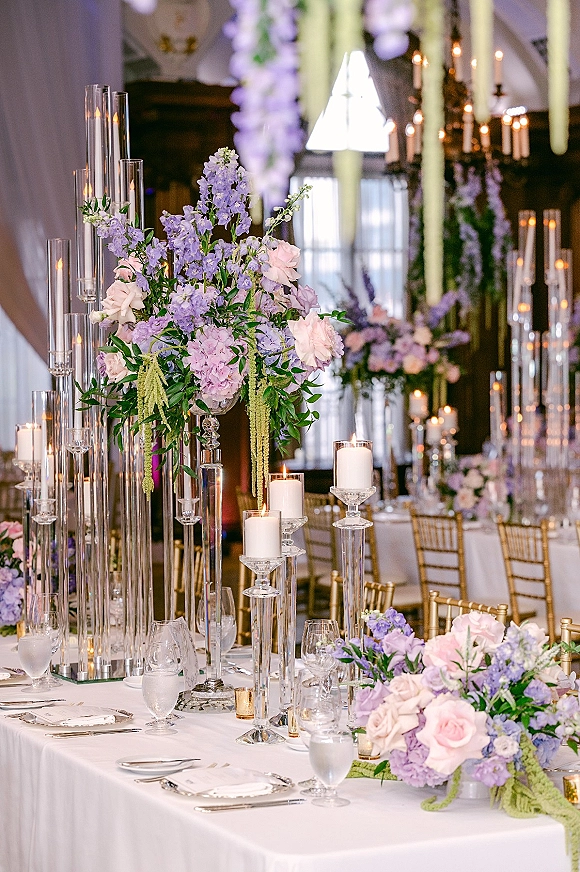 Reception tablescape with wedding table centerpieces in tall glass cylinder vases, taper candles, and gold Chiavari chairs beneath chandeliers