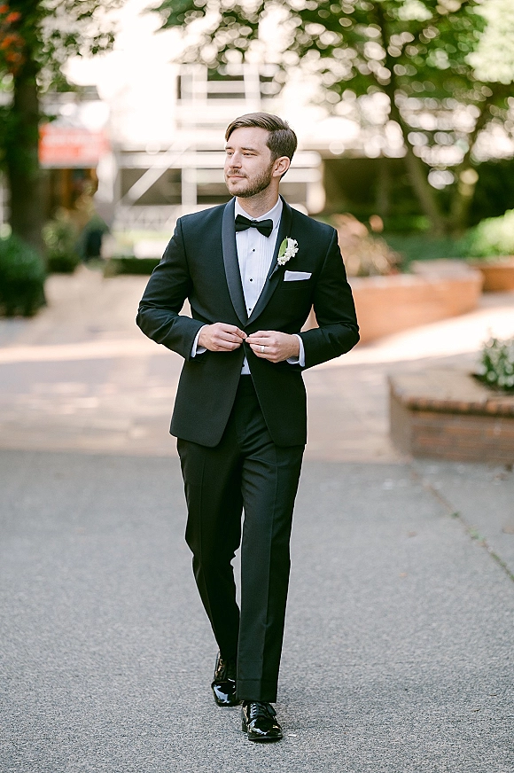Groom portrait in tuxedo walking along an outdoor walkway, wearing a black tuxedo with bow tie, boutonniere, and pocket square amid trees