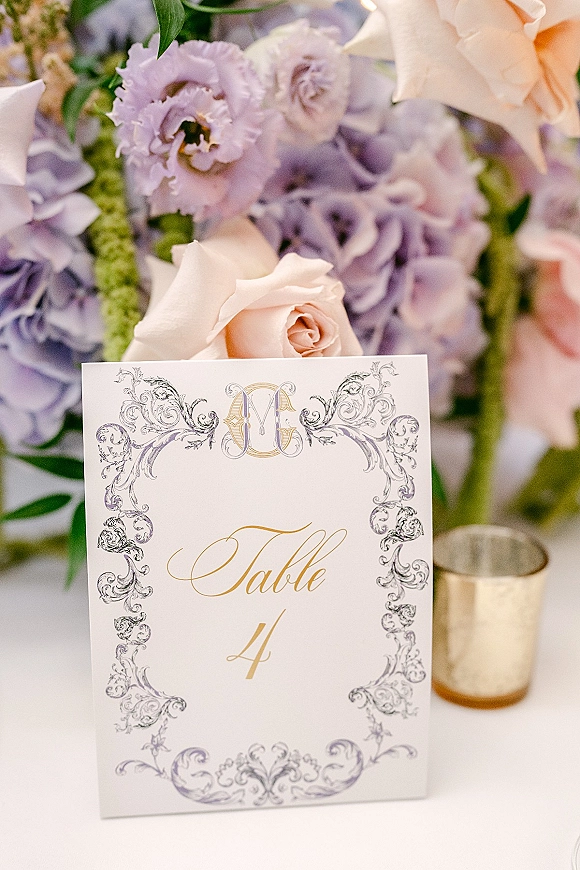 Wedding table number card with ornate monogram calligraphy, framed by roses and hydrangea centerpiece on a white tablecloth with candle votive