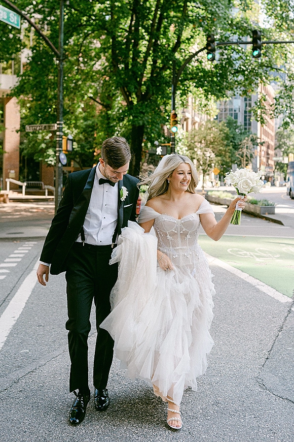 Couple portrait of bride and groom walking across a city street crosswalk, her tulle wedding dress and white bouquet beside his tuxedo