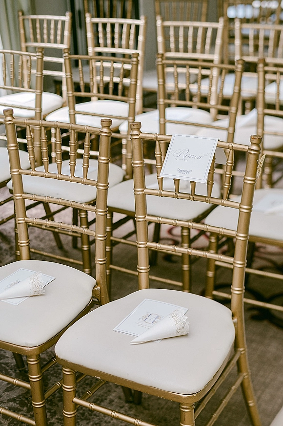 Ceremony seating with gold chiavari chairs and ivory cushions, reserved sign and programs on chairs, lace paper cones in indoor room