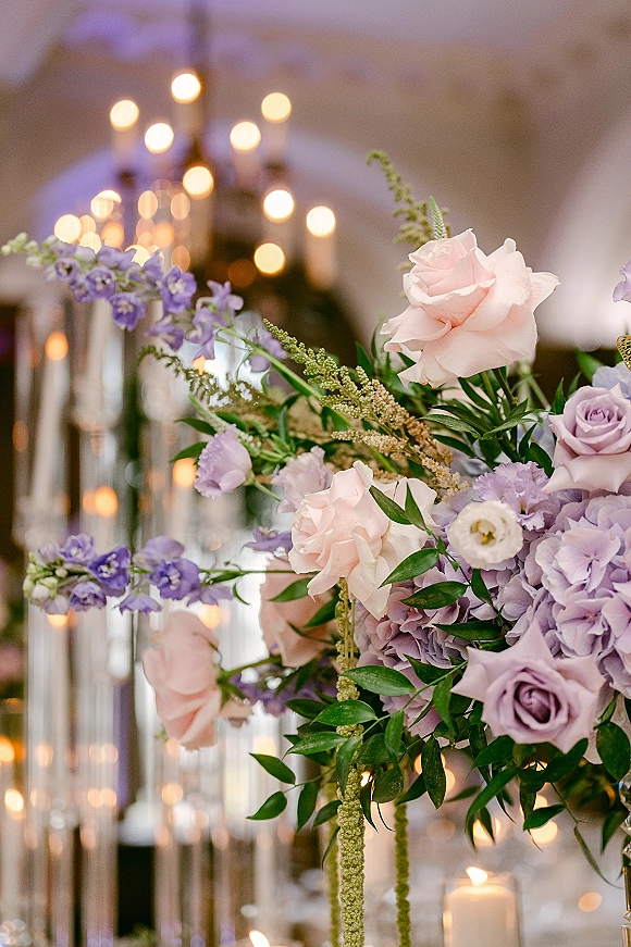 Wedding centerpiece with a tall floral centerpiece of roses, hydrangea and greenery, surrounded by pillar candles in glass holders and bokeh lights