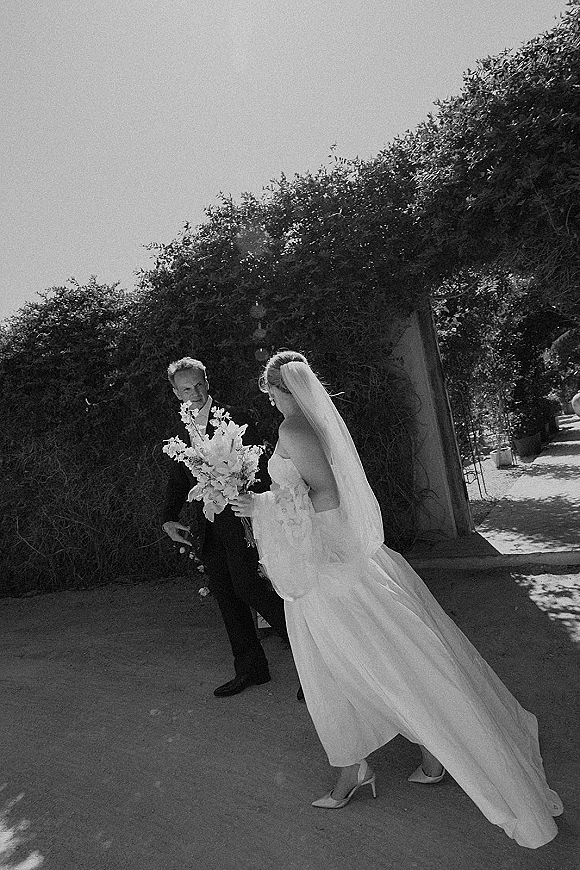 Wedding first look as bride walks to groom in a sunlit garden walkway, veil flowing, holding a bouquet, groom in tux by archway