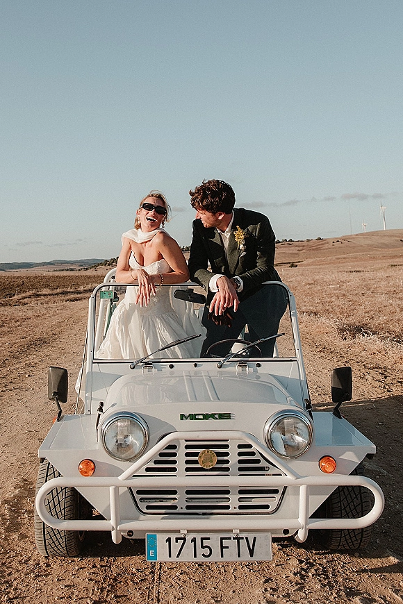 Couple portrait of bride and groom in car, bride laughing in sunglasses as he looks at her, with wind turbines on distant hills