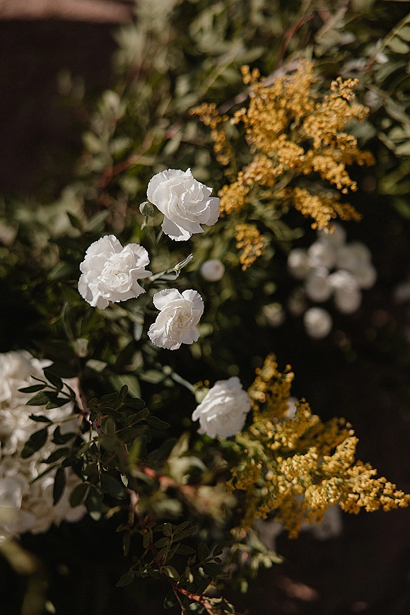 Wedding florals featuring white wedding flowers with yellow filler blooms and lush greenery, captured in natural light against blurred garden foliage