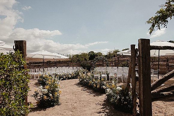 Outdoor ceremony setup with floral aisle arrangements leading to a circular flower arch, white chairs under umbrellas by rustic fencing