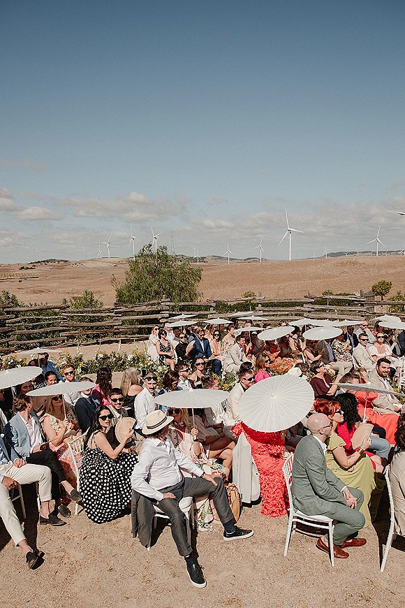Wedding guests at an outdoor wedding guests ceremony seated on white chairs, shading with paper parasols and fans under blue sky