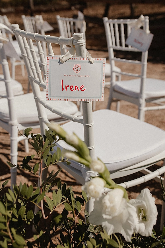 Wedding seating cards tied to white chiavari chairs with ribbon and floral greenery, sunlit outdoor ceremony seating in soft blur