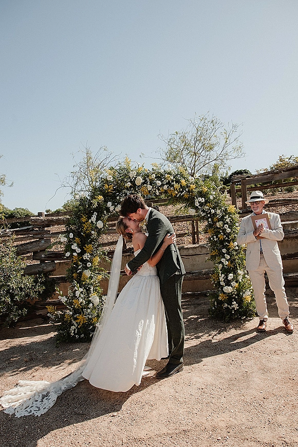 Wedding kiss under a round floral arch with yellow and white blooms, bride’s long veil and lace train flowing in sunny rustic outdoor setting