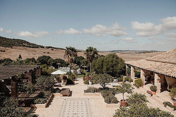 Outdoor wedding venue with palm trees, patio umbrellas, and lounge seating on a tiled terrace overlooking rolling hills and mountains