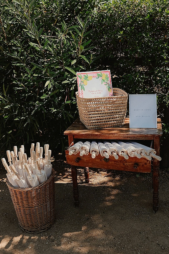 Wedding welcome table with a wedding welcome sign, wicker baskets of paper programs and parasols set on a wooden table by garden shrubs