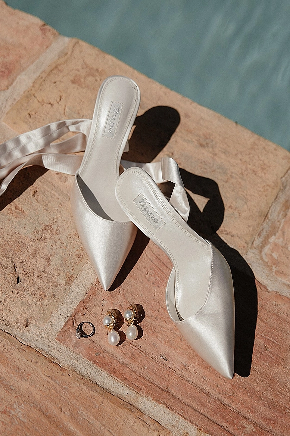 Bridal shoes in white satin wedding heels with ribbon ties beside pearl drop earrings and a ring on stone by water