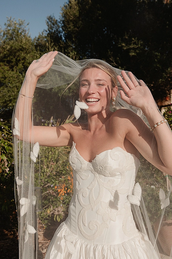 Bridal portrait of a bride holding veil, smiling in a strapless floral appliqué gown with butterfly veil, in a sunlit garden setting