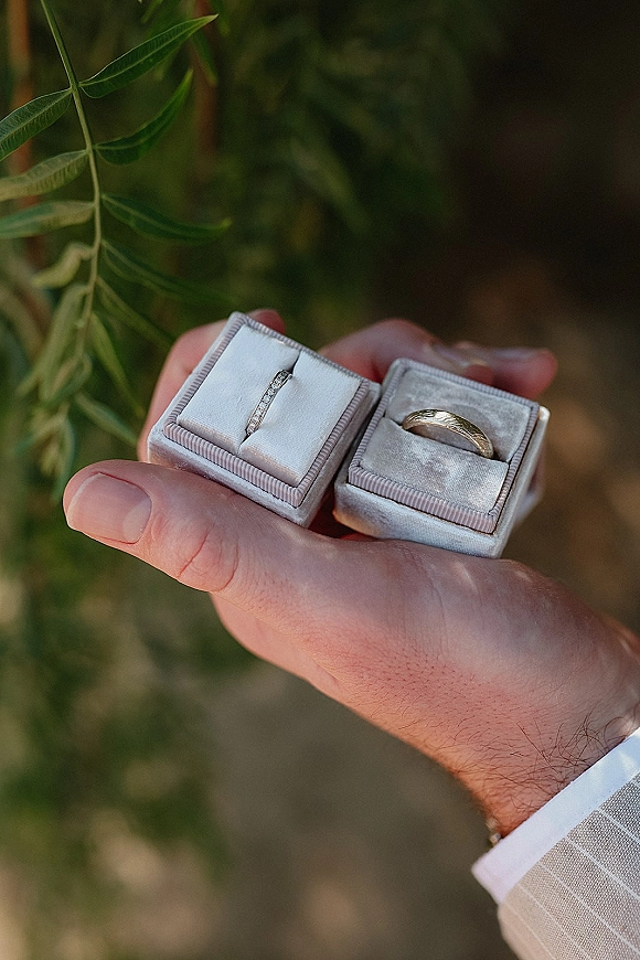 Wedding rings in a velvet ring box, diamond and gold bands held in a groom’s hand with suit sleeve against lush greenery backdrop