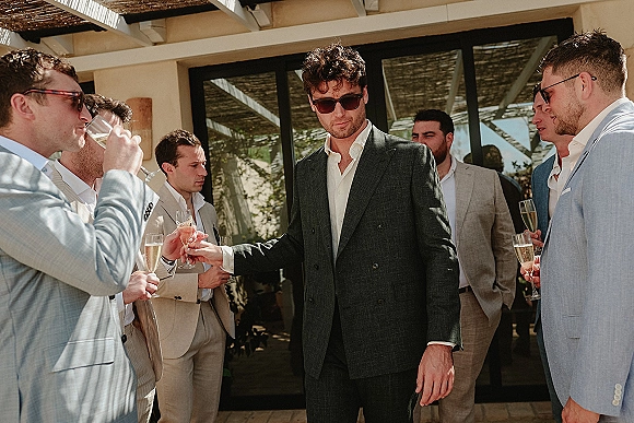 Groomsmen portrait of men in suits wearing sunglasses and holding champagne flutes on a sunny patio terrace by glass doors