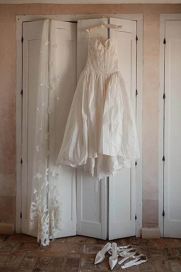 Wedding dress hanging on a hanger, strapless gown with lace bodice and tulle skirt, cathedral veil and ribbon-tied shoes by a white screen