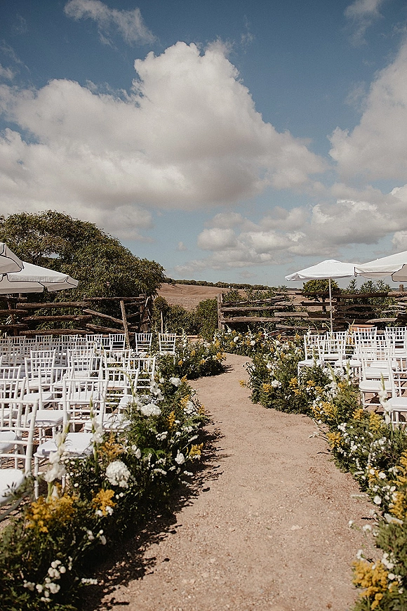 Ceremony aisle design with an outdoor ceremony aisle of white and yellow florals and greenery, lined by white chairs and patio umbrellas under blue sky