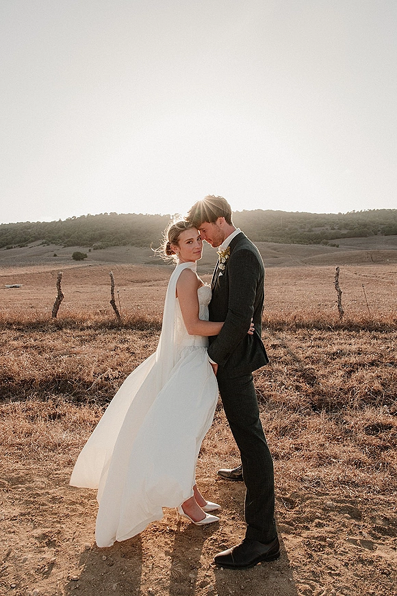 Couple portrait at sunset wedding portrait, bride in strapless dress with cape veil and heels, groom in dark suit, in a dry field by vineyard posts