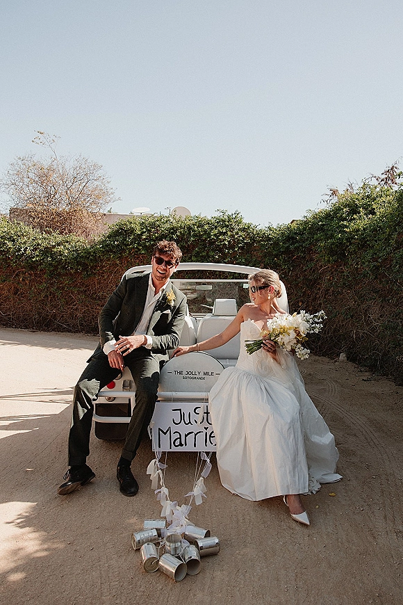 Couple portrait of bride and groom in sunglasses in a just married car, bouquet and veil visible, driving a convertible on a dirt road