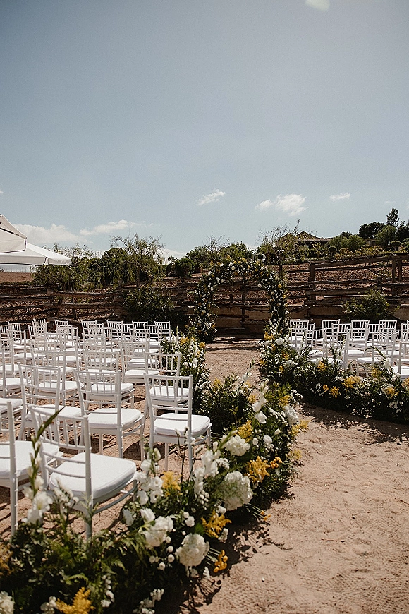 Ceremony setup with outdoor wedding ceremony seating of white Chiavari chairs facing a circular floral arch under a canopy on a hillside