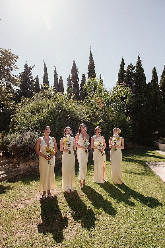 Bridesmaid portraits of a lineup in pale yellow dresses holding white and yellow bouquets on a sunny garden lawn by a stone wall