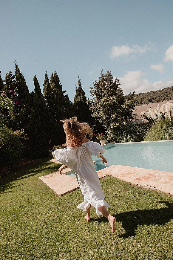 Flower girl moment as she runs barefoot in a white dress beside a villa pool, with lawn, stone deck, and hills under blue sky