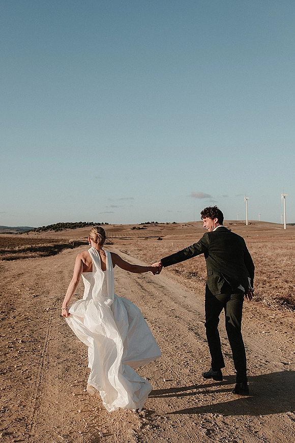 Couple portrait of bride and groom walking away holding hands down a dirt road, her backless dress and his black suit near wind turbines