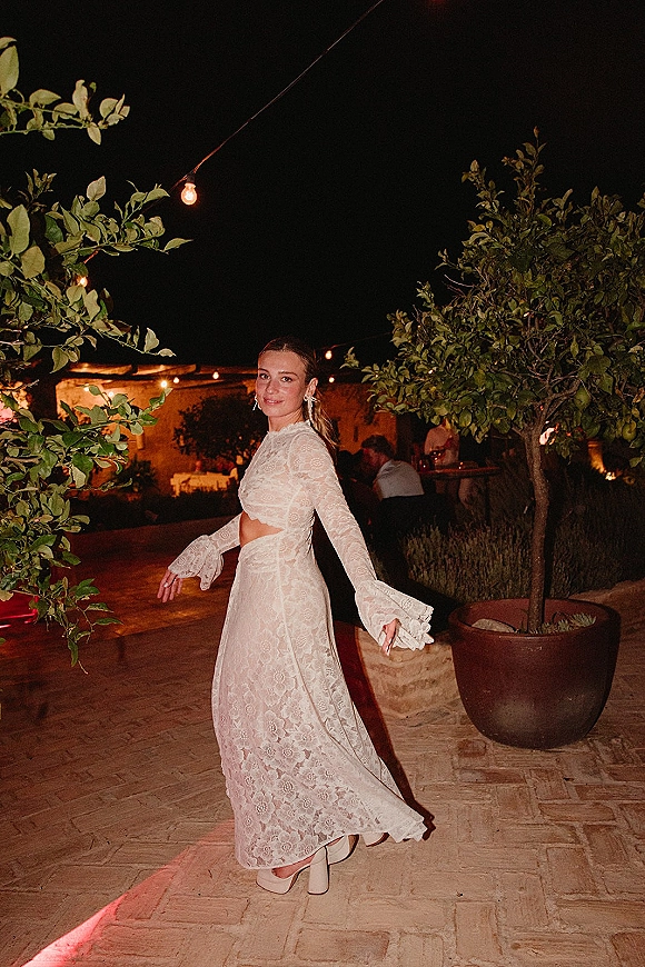 Bride portrait in a lace wedding dress with bell sleeves and midriff cutout, standing under string lights in an outdoor courtyard at night