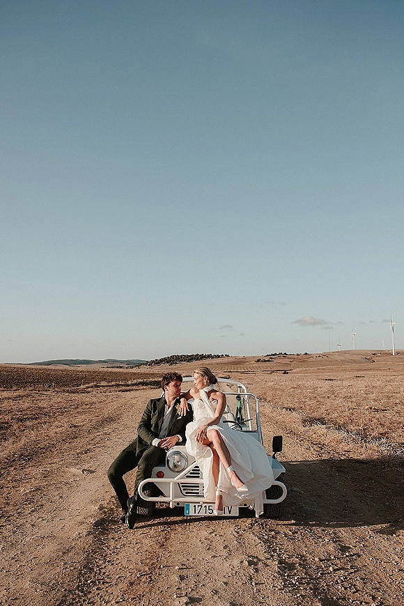 Couple portrait of bride in veil and dress sitting on a vintage car hood as groom in suit leans beside her on a dirt road with wind turbines