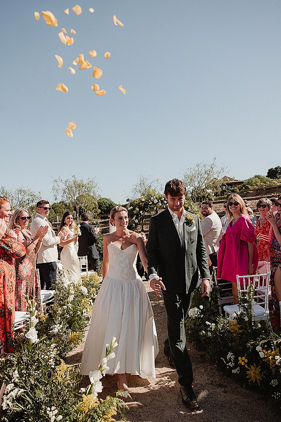 Wedding recessional as bride and groom walk the aisle holding hands, flower petal toss in the air, with white chairs in a sunny garden setting