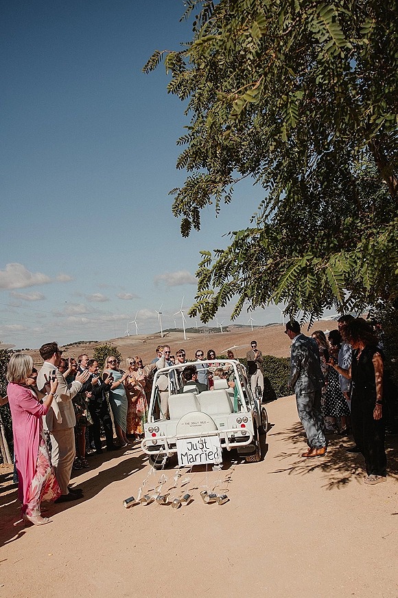 Wedding send off with a white just married car sign and tin can streamers as guests line a dirt road under trees and blue sky