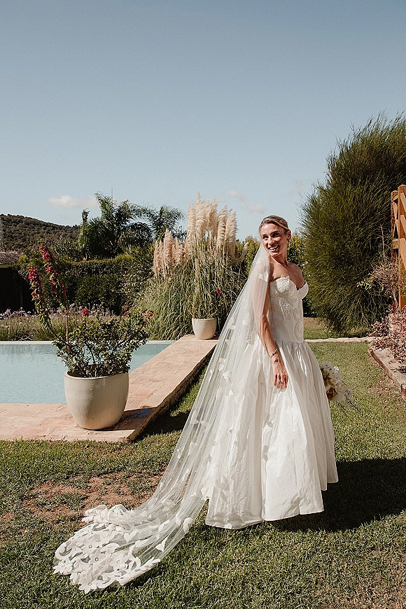 Bridal portrait of a smiling bride in a strapless wedding dress with long lace appliqué veil, standing poolside on a garden lawn under blue sky