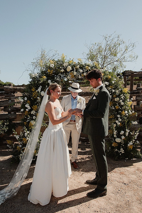 Wedding vows as bride in strapless dress and veil faces groom in suit under a white and yellow floral arch by a rustic fence in desert sun