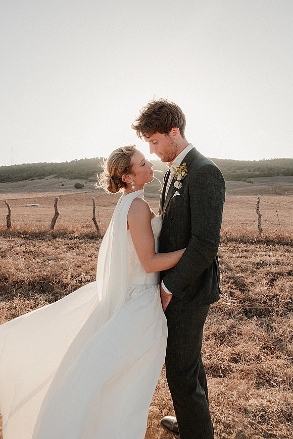 Couple portrait of bride and groom embracing, her long veil blowing in wind, in an open field with hills and fence posts behind them