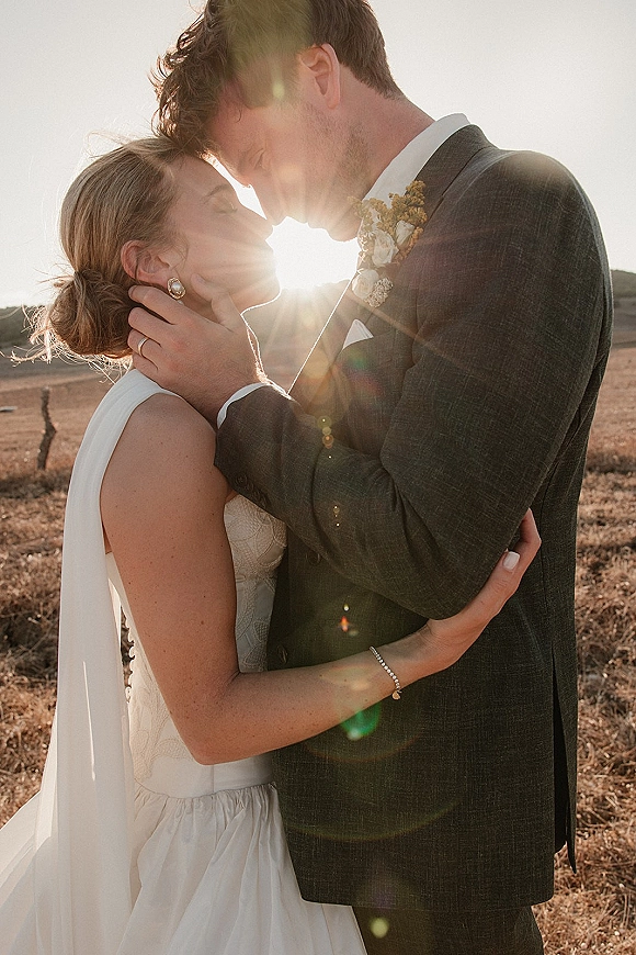 Wedding kiss portrait at sunset as the bride in a veil holds the groom’s face, backlit in an open field with dry hills behind