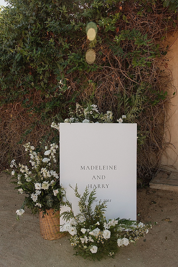 Wedding welcome sign with black typography on a white board, framed by white flowers and greenery against an ivy wall backdrop