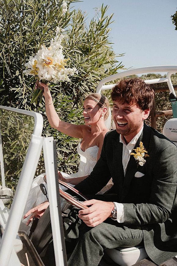 Wedding getaway moment as bride in veil holds bouquet beside laughing groom on a golf cart, riding past green hedges under blue sky