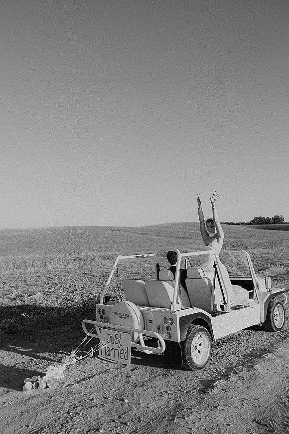 Wedding getaway car with a just married car sign and tin cans, bride in wedding dress leaning from the convertible on a dirt road field