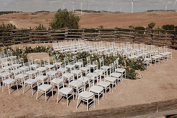 Ceremony setup with outdoor ceremony seating of white chairs lining a floral-bordered aisle, greenery garland by a rustic fence and hills