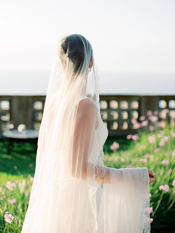 Bridal portrait of a bride from behind in a long wedding veil and lace long-sleeve dress, by garden flowers with an ocean view beyond